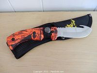 Side view of the Elk Ridge hunting knife with orange and black wood grain handle resting on a black nylon sheath with yellow logo.