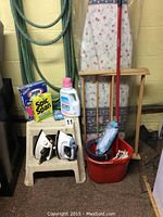 Photo showing ironing board, two electric irons on step stool, garden hose on wall, red pail with mop and cleaning cloths, several cleaning products including Spic and Span, Clorox, and Zero detergent.
