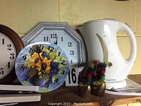 Three battery operated wall clocks including round wooden framed clock, silver octagonal framed clock, and floral sunflower-themed clock with battery operated mechanism visible.