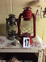 Two oil lamps on floral tablecloth. One red standing 12 inches tall marked 'JUNIOR', one silver 10 inches tall marked 'Feuerhand Hansa Lux'. Extra wick material rolled on table.