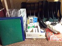 Photo shows black and white plastic magazine holders, blue and green binders, Texas Instruments adding machine calculator, box of index cards, hole punch, stapler, label box, and a lap computer table with colorful bicycle design.