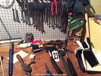 Wide view of various hand tools on a pegboard wall and table including wrenches, pliers, vice grips, hand drill, and brush.