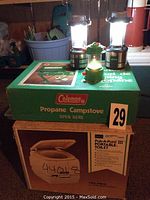 Photo showing two North 49 battery operated lanterns placed on a green Coleman propane camp stove box, and Pak-A-Pot III portable toilet box below.