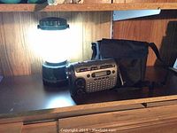Photo shows a lit 13 inch battery lantern beside a Sylvania wind-up AM/FM radio with flashlight and black carrying case on a wooden surface.