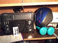 Photo displaying a Venturer 2-way short wave radio with multiple controls, a blue bike helmet, a black hockey helmet, and two teal dumbbells marked 1kg each on a wooden shelf.