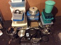 Overall view of kitchenware lot including pans, pressure cooker, bakeware, and plastic containers stacked and arranged on a surface