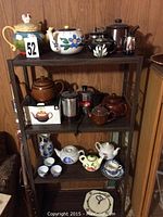 Photo showing a variety of tea pots and cups displayed on a three-tier black metal shelf against a wood-paneled wall.