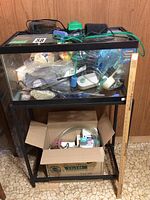 Full view of 57L glass fish tank on black metal stand with accessories inside the tank and a cardboard box filled with aquarium supplies underneath.