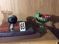 Wood-paneled shelf showing two black lacquer Asian spheres with floral designs and carnival glass compote with fruit decoration.