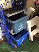 Recycle center setup showing blue metal rack holding two blue plastic recycle bins with white recycle symbols, white two-step plastic stool beside it, and large beige garbage can with lid behind.