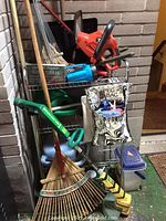 Photo of yard implements including trimmers, rakes, peg bag with clothespins, and knee pads organized on a small metal rack near brick wall.