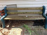 Photograph of outdoor bench against a siding wall showing wood slats and green plastic frame, situated on patio tiles with leaves and vegetation around.