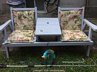 Front view of gray wooden 2-seater bench with two floral cushions on backrest and seat, center table area, and two metal rods with fish-shaped watering attachments below.