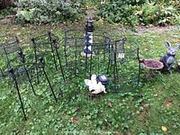 Six black iron plant stands of varying heights along with several garden figurines and a small iron bird bath placed on grass outdoors.