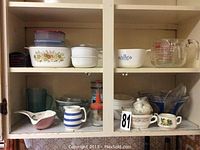 Photo showing the kitchen cabinet shelf containing assorted Pyrex, Corningware and Fire King kitchenware items including casserole dishes, bowls, measuring cups, mugs, pitchers and small containers.