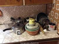 Photo showing kitchenware including metal pots, ceramic bowls stacked, yellow measuring cups nested inside bowls, two black frying pans, glass measuring cup, wooden rolling pin, and other kitchen utensils.