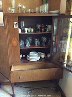 Wooden pedestal china cabinet with glass door open, showing stacked dishes and glassware on shelves and drawer below.