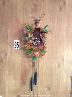 Front view of wooden mechanical cuckoo clock mounted on wooden wall, showing deer head, bird, leaves, pinecone weights, and pendulum.