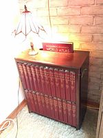 Wooden bookcase filled with 24-volume Encyclopedia Britannica set and the 1960 World Atlas on top, with stained glass lamp on the corner.