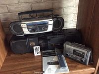 Photo of three portable radios on a wood shelf: Sylvania duo cassette player with AM/FM radio and detachable speakers at the back; Electorhome wind-up radio on right; Sharp QT-CD33H CD AM/FM radio with cassette player on left with visible manual and a numbered lot marker.