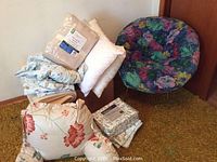 Photo of bed sheet sets, blankets, retro floral chair, and cushions arranged on carpeted floor