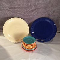 Two large Fiesta Ware chargers in ivory and cobalt blue glaze with a stack of eight small plates in bright colors including orange, yellow, turquoise, and red, one turquoise marked 'Frolic England'.