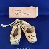 Pair of worn pink satin pointe ballet shoes laid flat with attached ribbons and original signed box visible above.
