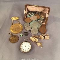 Overview of all lot contents including coins spilling from case, pocket watch at center, cufflinks, medals, ring, charm