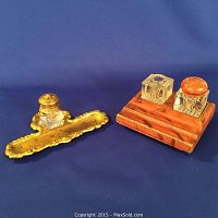 Two antique inkwells displayed on blue surface; one with ornately detailed ormolu brass base and pressed glass bottle, another with two glass bottles on wooden painted base.