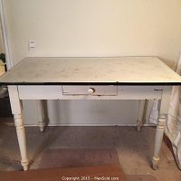 Front view of vintage kitchen table showing white enamel top with black trim and wood turned legs. Drawer front visible with knob.