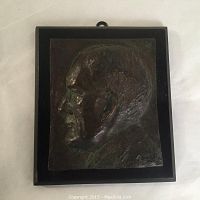 Front view of bronze plaque depicting a man's side profile in relief, mounted on a black wooden base.