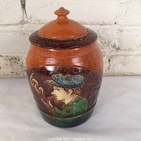 Front view showing man smoking pipe in majolica relief design on jar with lid on top.