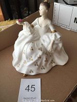 Front and side view of Royal Doulton My Love figurine held by seated woman with gold-decorated white dress and pink flower in hand.