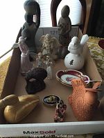 Photo showing assortment of decorative figurines including angels, birds, and carved stone statues in a cardboard box