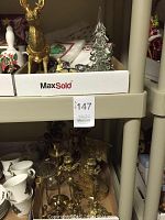 Photo of the lot contents on shelving showing assorted brass pieces and Christmas decorations including brass reindeer, glass Christmas trees, brass candlesticks, and napkin rings.