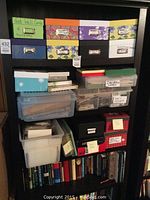 View of shelf with labeled photo boxes containing cards and some unseen content in plastic containers, along with books on lower shelves.