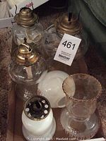 Photo showing a box containing six glass oil lamps and chimneys with metal fittings, some with brass tops and wick adjusters.