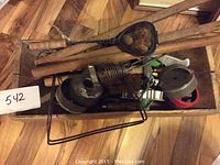 Top view of wooden box containing antique kitchen implements including a metal perforated scoop, wooden handles and other metal utensils.