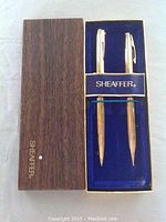 Two gold-tone Sheaffer pens inside blue velvet-lined wooden box with Sheaffer branding on box lid and on pen holder strap.