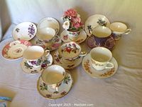 Seven sets of porcelain tea cups with matching saucers in different floral patterns arranged on a white cloth background with an additional porcelain vase with artificial flowers.