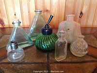 Seven vintage glass perfume bottles arranged on a table in front of a wooden wall. Variation in shapes, some with stoppers and atomizers.
