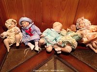 Full view of all five Ashton Drake Galleries dolls arranged seated on wood corner shelf, showing clothing and facial features.