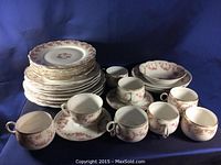 Full display of various plates, cups, saucers, and bowls with bridal rose pattern on white porcelain with gold trim. Multiple stacks of plates shown with cups and saucers in foreground.