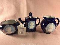 Front view of the three-piece set showing blue ceramic bowl, square teapot, and pitcher, each with white silhouette medallion and gold trim.