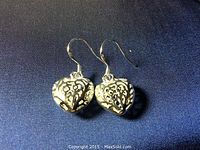 Close-up photo of a pair of sterling silver earrings shaped like hearts with intricate filigree design on a dark fabric background.