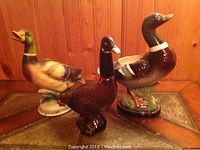 Wide shot of three goose figurines (two ceramic, one glass) on a tiled surface with wood-paneled background