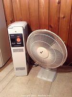 DeLonghi space heater positioned next to a white oscillating fan, both placed against a wood-paneled wall