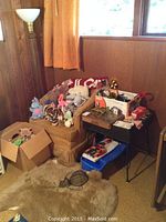 Full view showing the wicker upholstered chair with multiple stuffed animals on it, a cardboard box of stuffed animals on the floor, the black game table filled with boxes, and the brass floor lamp behind the chair.