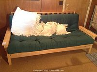 Futon sofa bed with light wood frame and green cushioned mattress, includes white fringe cushions and blanket visible on couch.