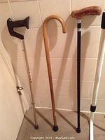 Three walking canes and white metal bed safety rail with straps against tiled wall and brown carpeted floor.
